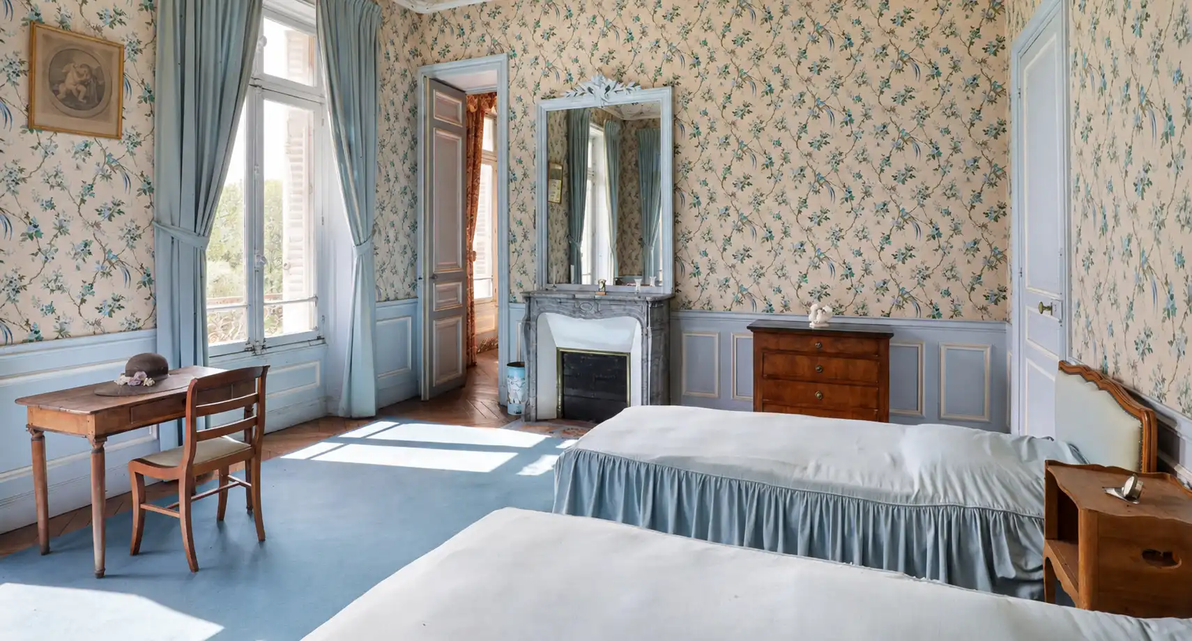 Chambre ancienne aux murs et rideaux bleu à fleurs, deux lits, bureau et cheminée à miroir