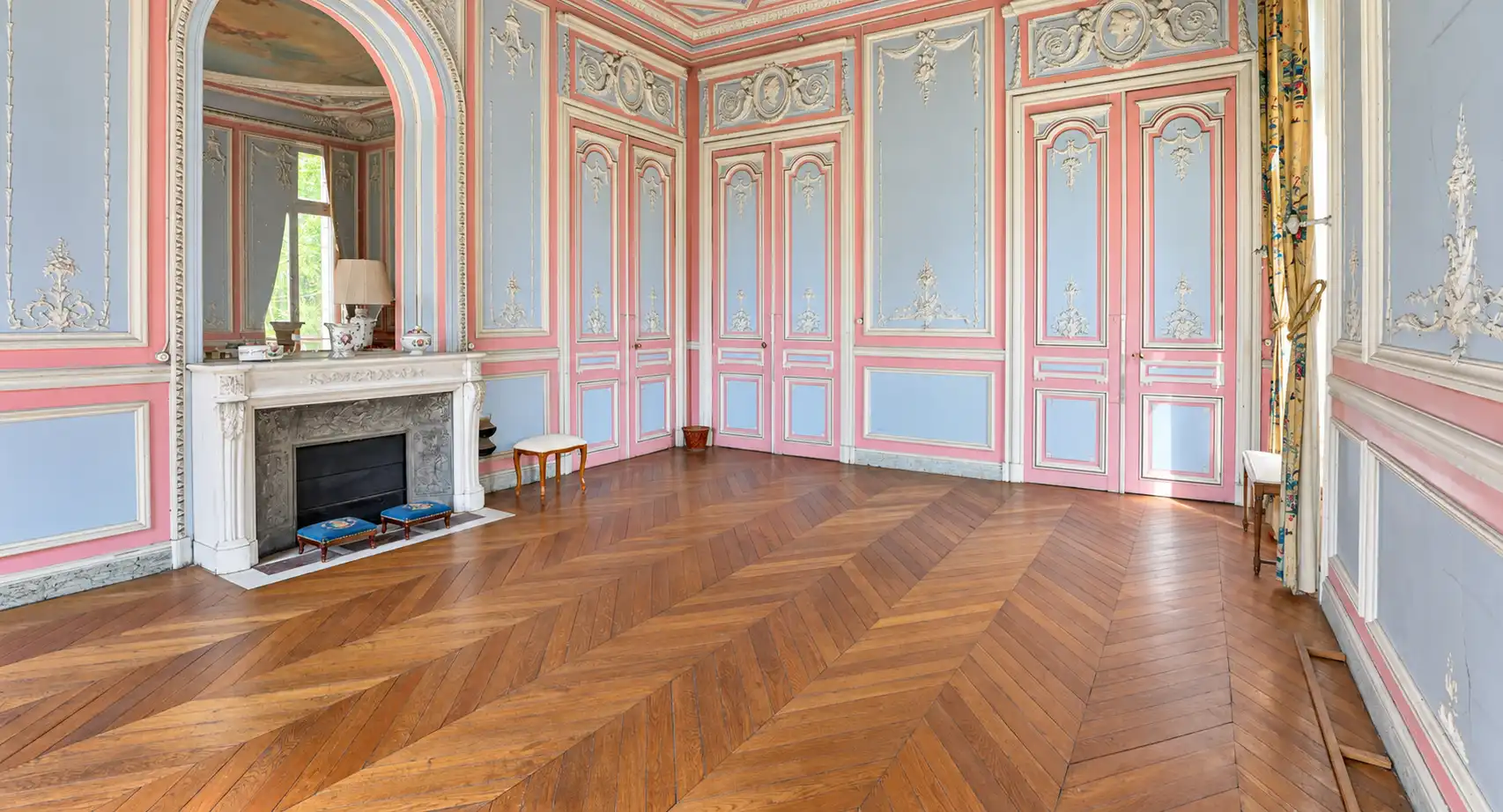 Salon vide aux boiseries bleues et roses, cheminée en marbre blanc, parquet en chevrons