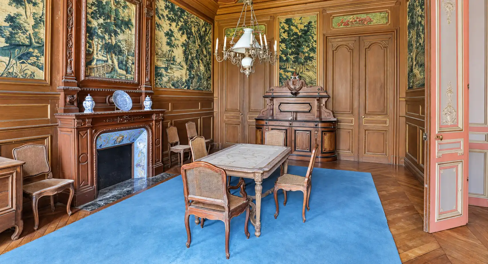 Salle à manger lambrissée de chêne avec tapisseries de verdure, table et chaises anciennes sur tapis bleu