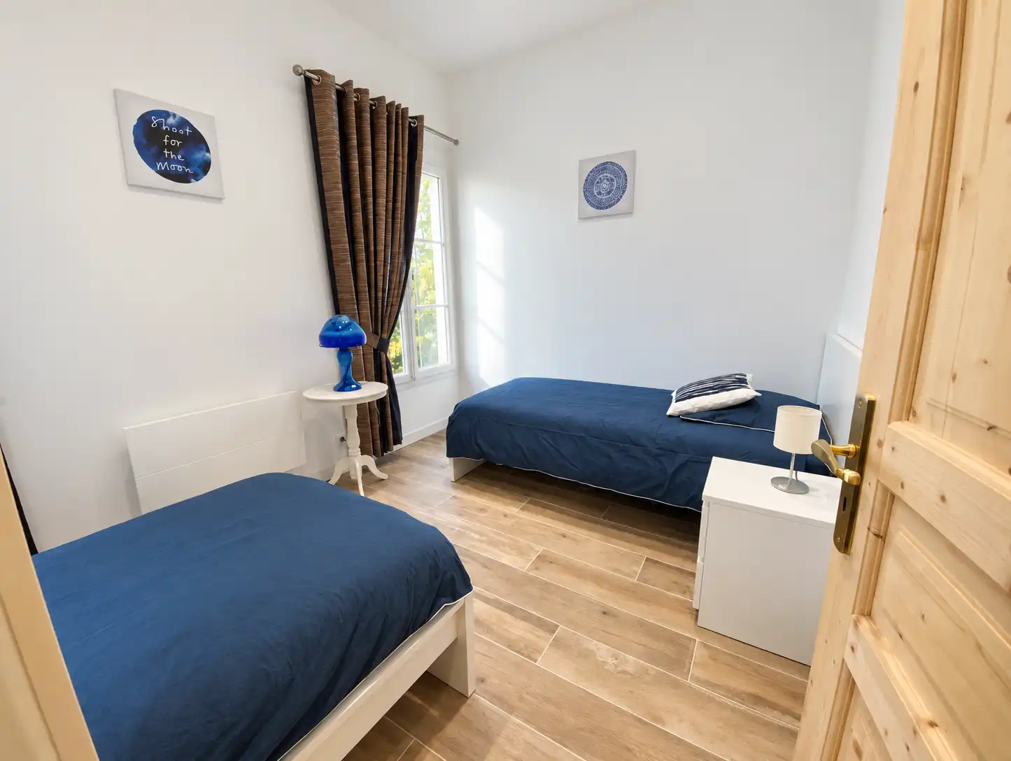 Deux lits simples aux couvre-lits bleus, rideaux bruns et parquet clair, au Monarque
