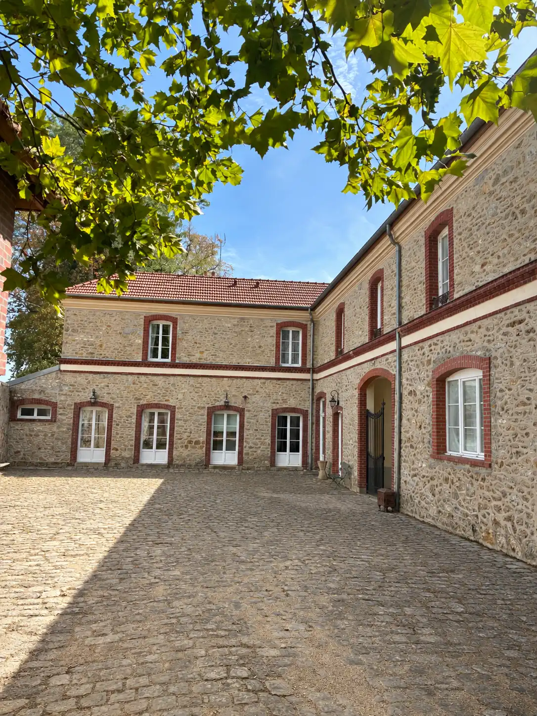 Cour pavée et bâtiments en pierre meulière vus sous les feuilles d'un platane